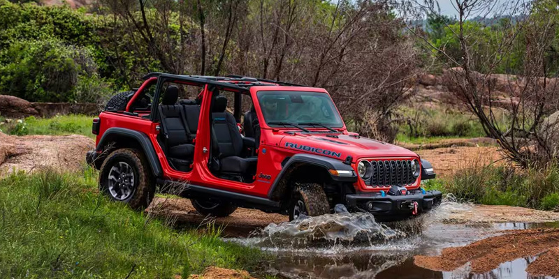A front three-quarter view of a red Jeep Wrangler Rubicon with its doors and roof removed, driving through a muddy water puddle in an off-road terrain.