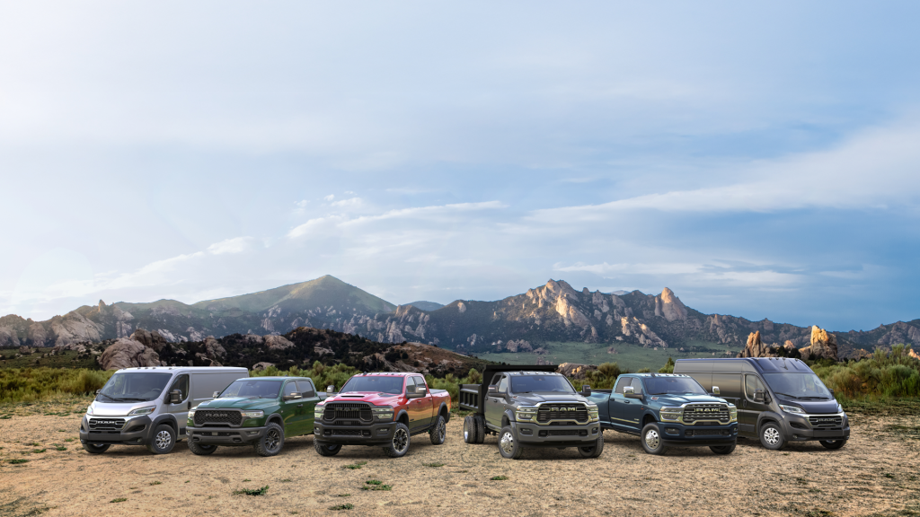 A lineup of various RAM vehicles, including pickup trucks and cargo vans in different colors, parked in a row on a dirt field with a scenic mountain range under a cloudy sky in the background.